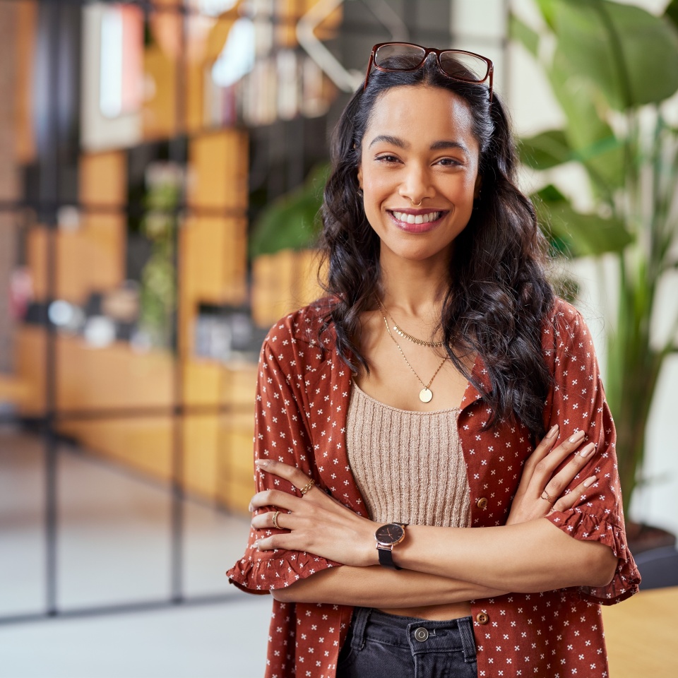 Portrait of smiling young multiethnic woman looking at camera with crossed arms. Successful latin business woman standing in modern office with copy space. Young university hispanic girl with smile.