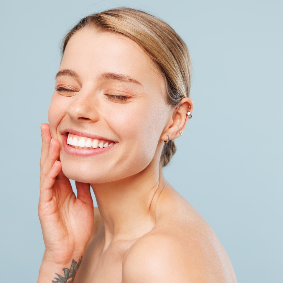 Close up happy half naked topless young woman 20s with nude make up close eyes with toothy smile touch face isolated on pastel blue color background. Skin care healthcare cosmetic procedures concept
