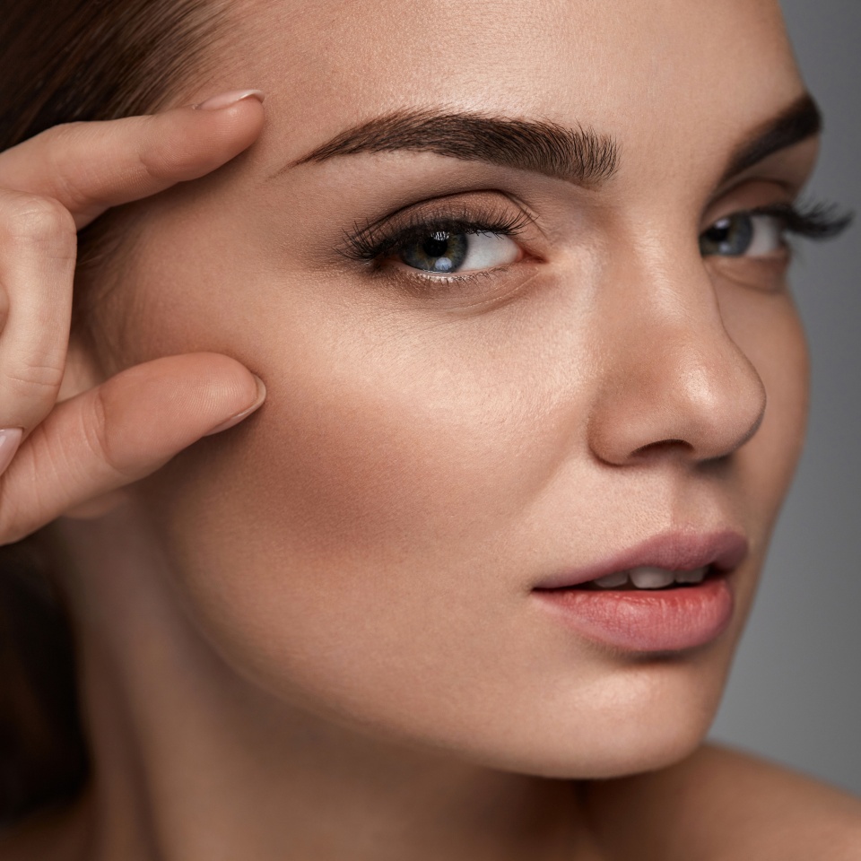 Woman Beauty Face. Portrait Of Beautiful Young Woman With Professional Makeup And Perfect Smooth Healthy Skin. Sexy Female Model With Fingers Near Face On Grey Background. Skin Care. High Resolution